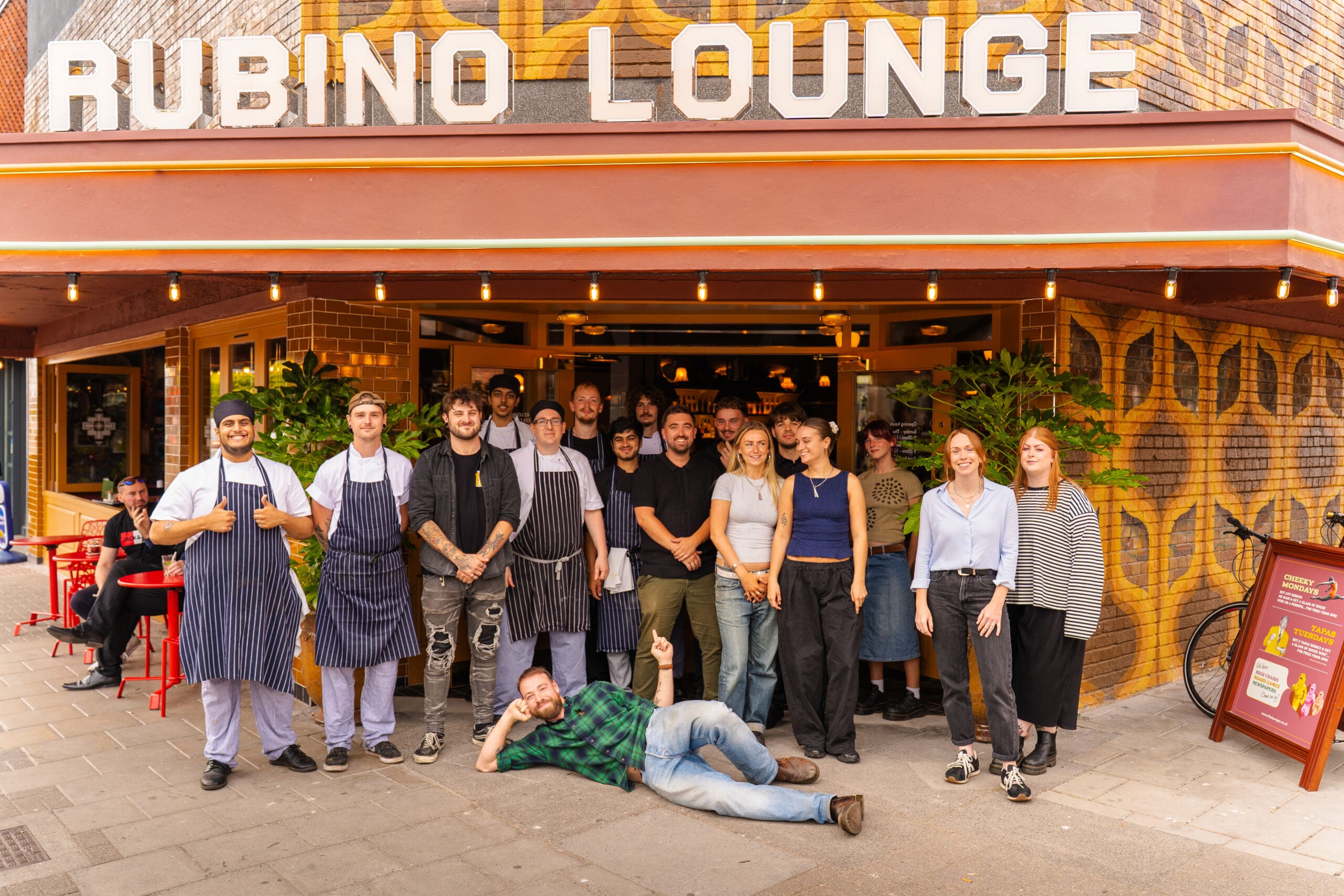 A group of people, some in aprons, pose and smile in front of Rubino Lounge, a restaurant with bold signage. One person lies playfully on the ground near the espino bushes by the outdoor tables visible to the side.