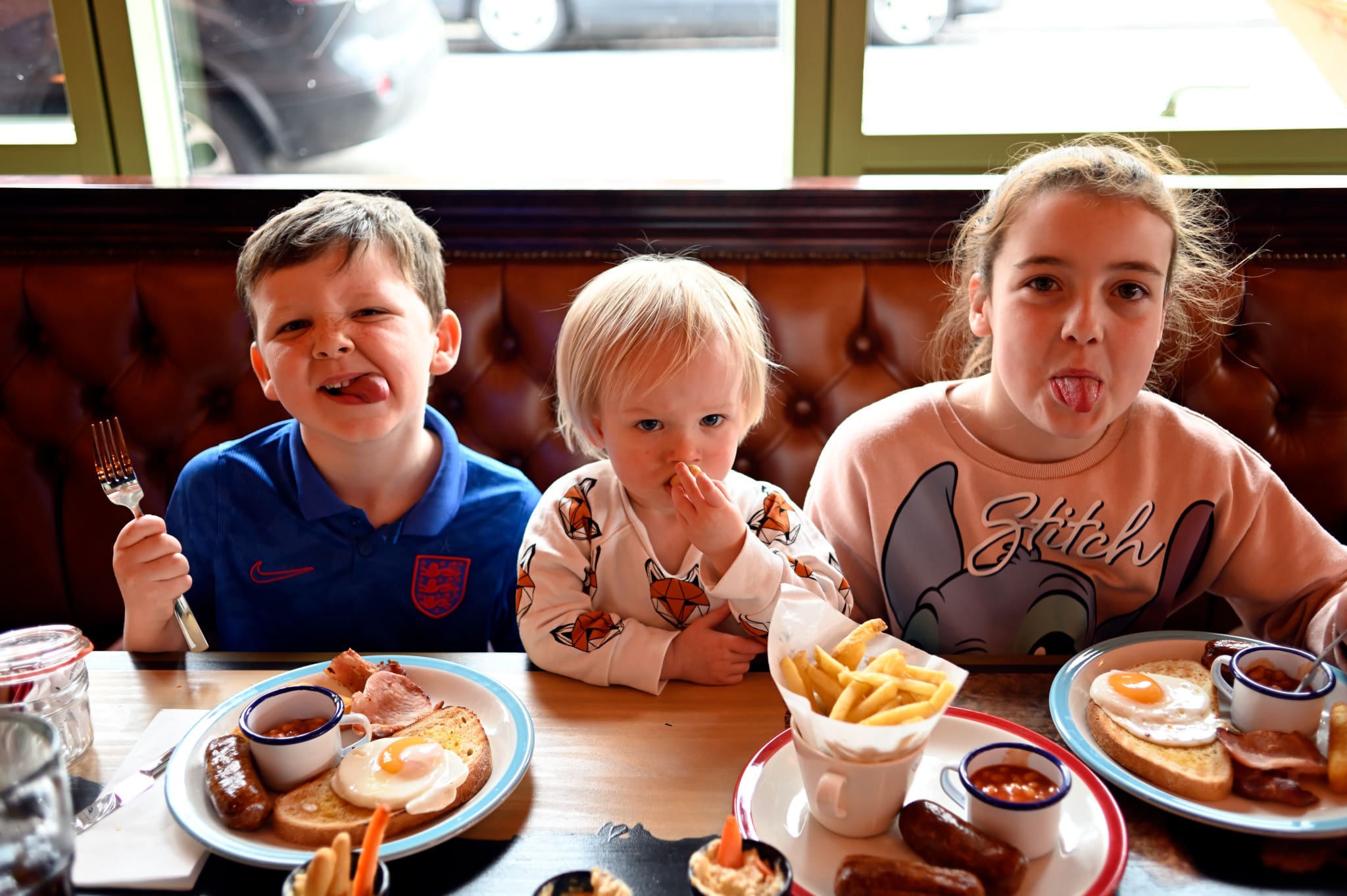 Three children sit at a table with plates of breakfast food, including fries, sausages, eggs, and beans. The two older kids are sticking out their tongues, while the youngest looks at the camera, finger in mouth.