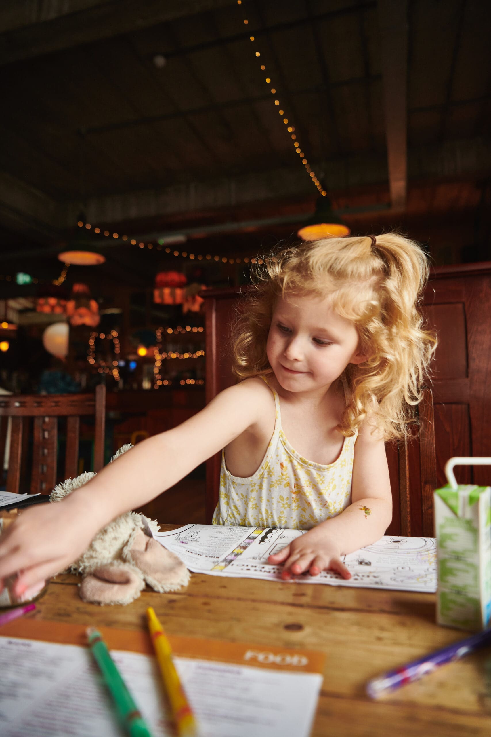 A young girl with curly blonde hair sits at a wooden table in a warmly lit restaurant, reaching for crayons while coloring on a placemat. A stuffed animal and juice box are on the table beside her.