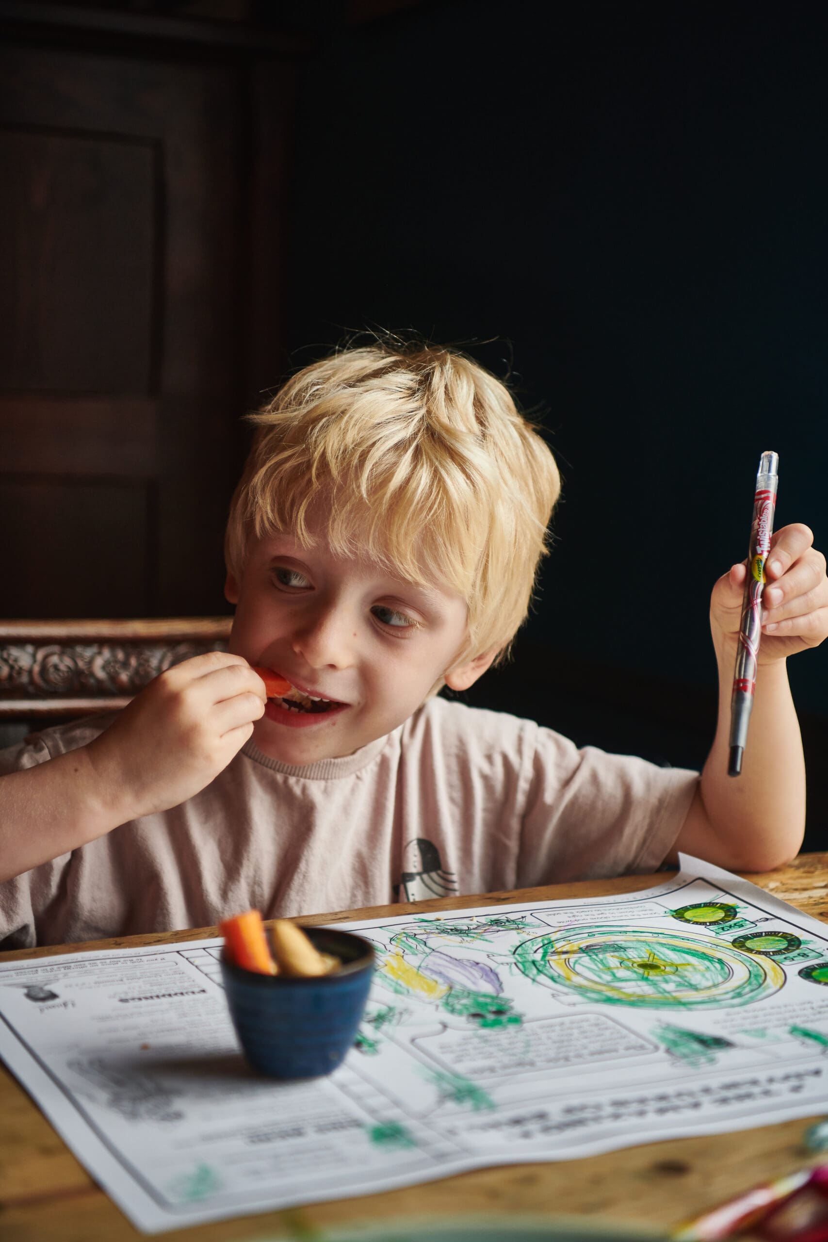 A young blond boy sits at a table, holding a colored pen in one hand and eating carrot sticks with the other. In front of him is a partially colored placemat with green and yellow drawings.