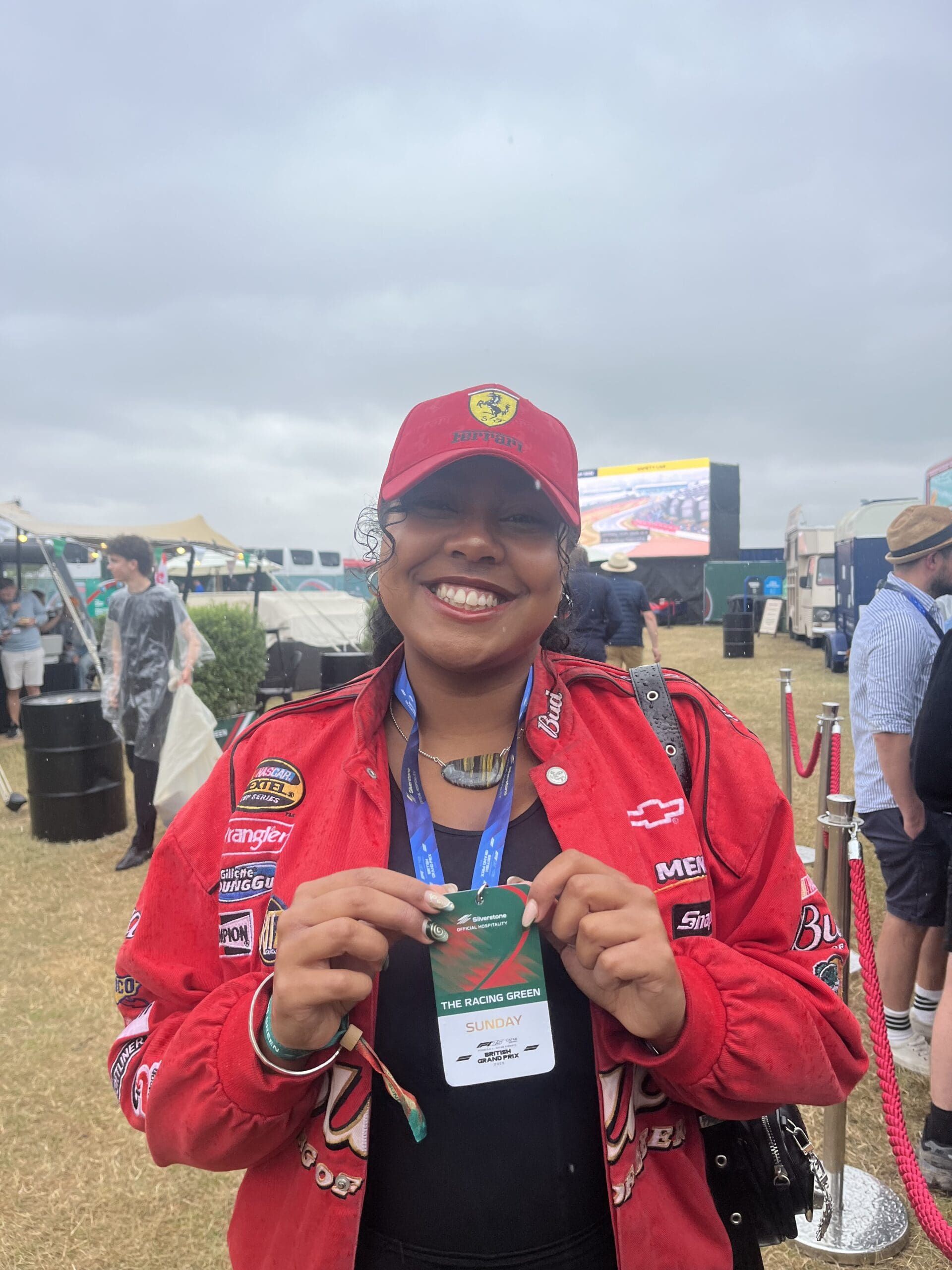 A smiling person wearing a red Ferrari cap and racing jacket holds up a green event pass. They are outdoors at a motorsport event, with other attendees and a large screen visible in the background under cloudy skies.