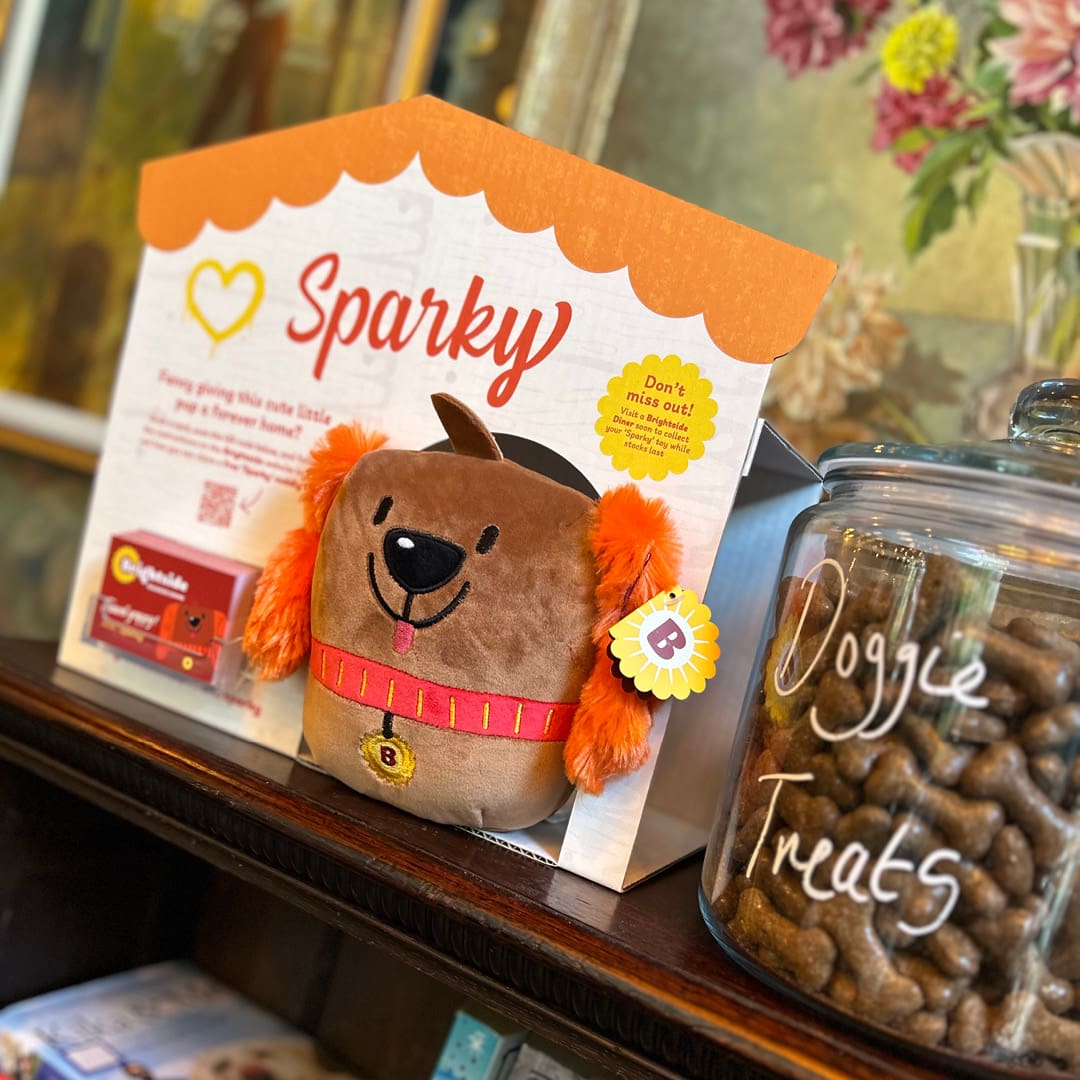 A plush dog toy with orange ears labeled "Sparky" is displayed in a house-shaped box next to a glass jar of dog treats labeled "Doggie Treats" on a wooden shelf.