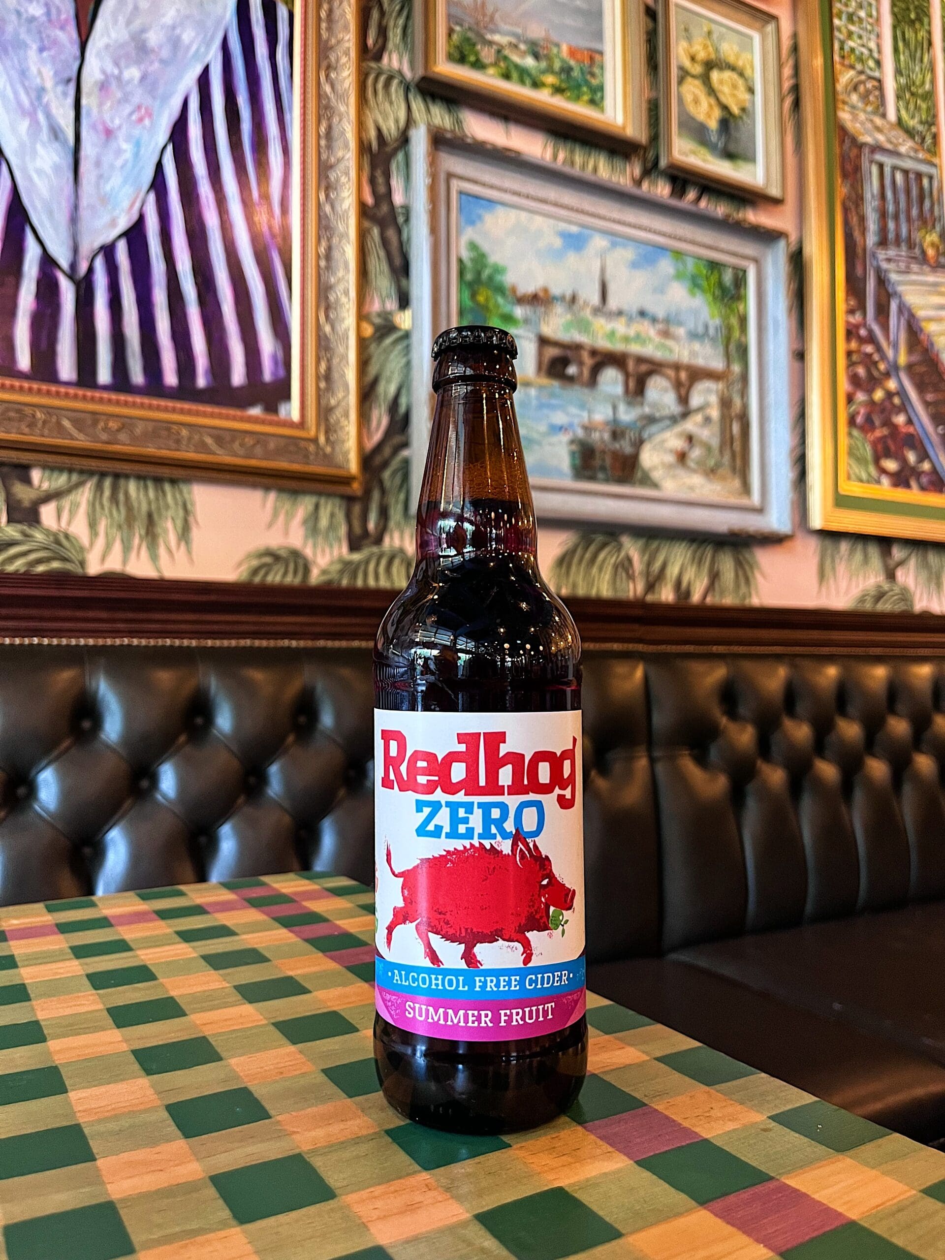 A bottle of Redhog Zero alcohol-free summer fruit cider sits on a checkered table in a stylish cafe with a tufted leather bench and framed artwork on the walls in the background.