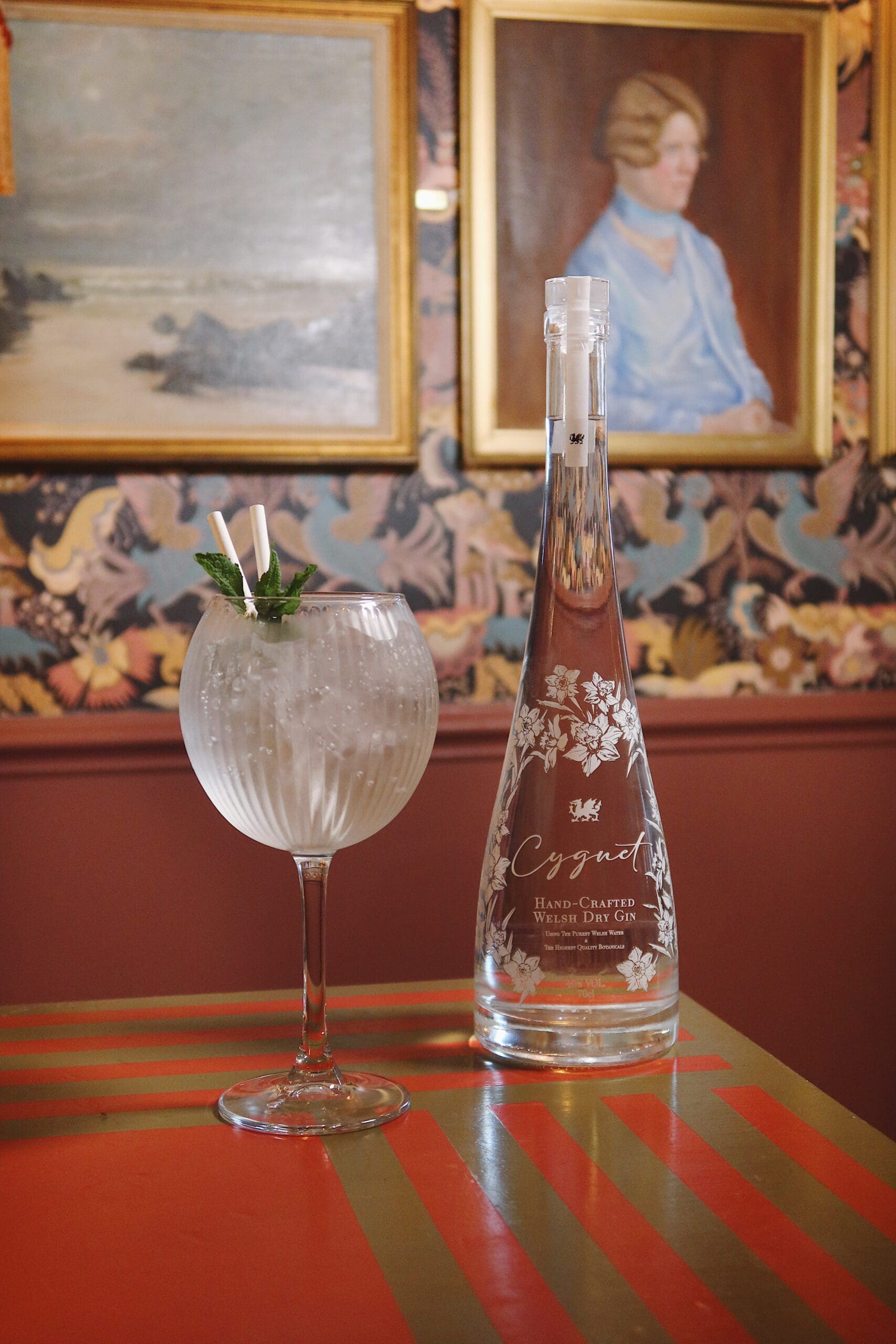 A large glass of clear cocktail with ice, mint garnish, and two straws sits next to a tall, ornate glass bottle on a striped table. Floral wallpaper and framed portraits are visible in the background.