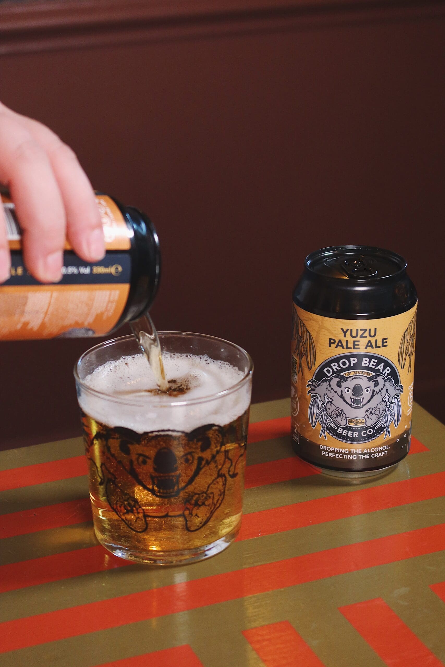 A hand pours Yuzu Pale Ale from a can into a glass with a koala design. Another can of the same beer sits nearby on a striped surface.