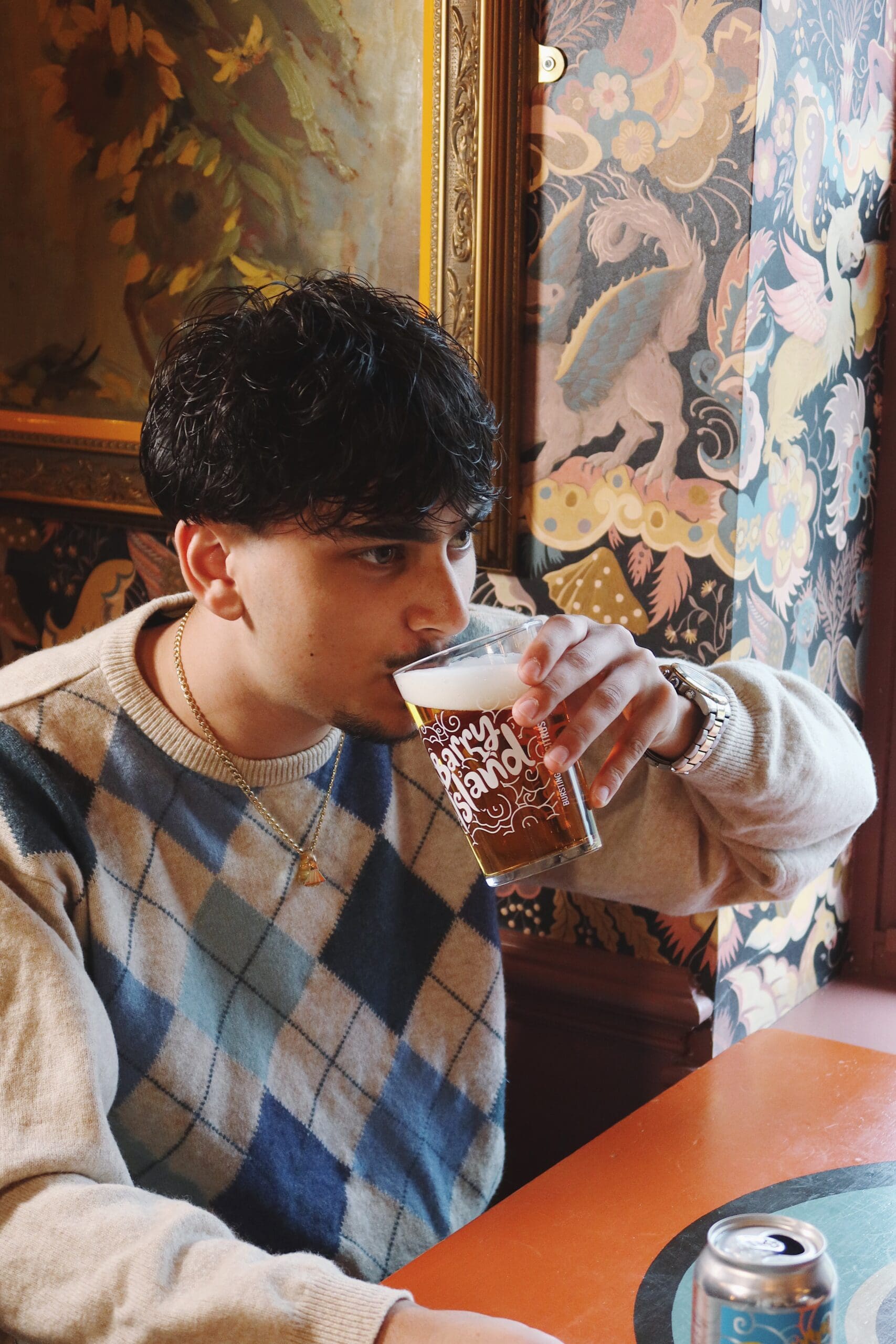 A young man with dark, wavy hair sips beer from a glass while sitting at a table. He wears an argyle sweater and a gold necklace. The background features ornate, colorful wallpaper with floral and dragon designs.