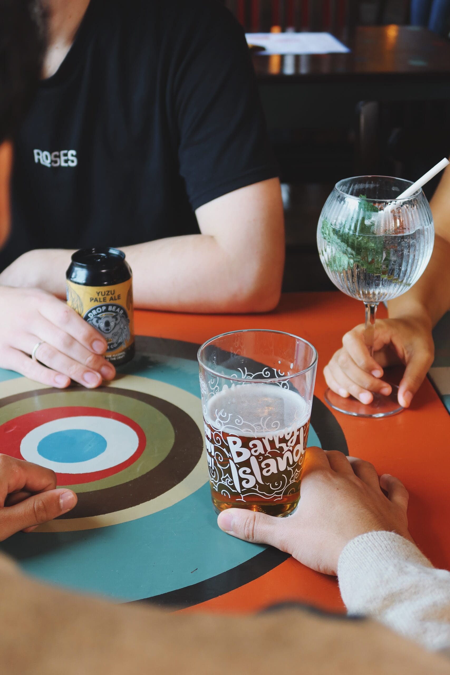 Three people sit around a table with drinks: a beer in a Barley Island glass, a can, and a cocktail with a straw and mint. Their hands and arms are visible, but their faces are out of frame.