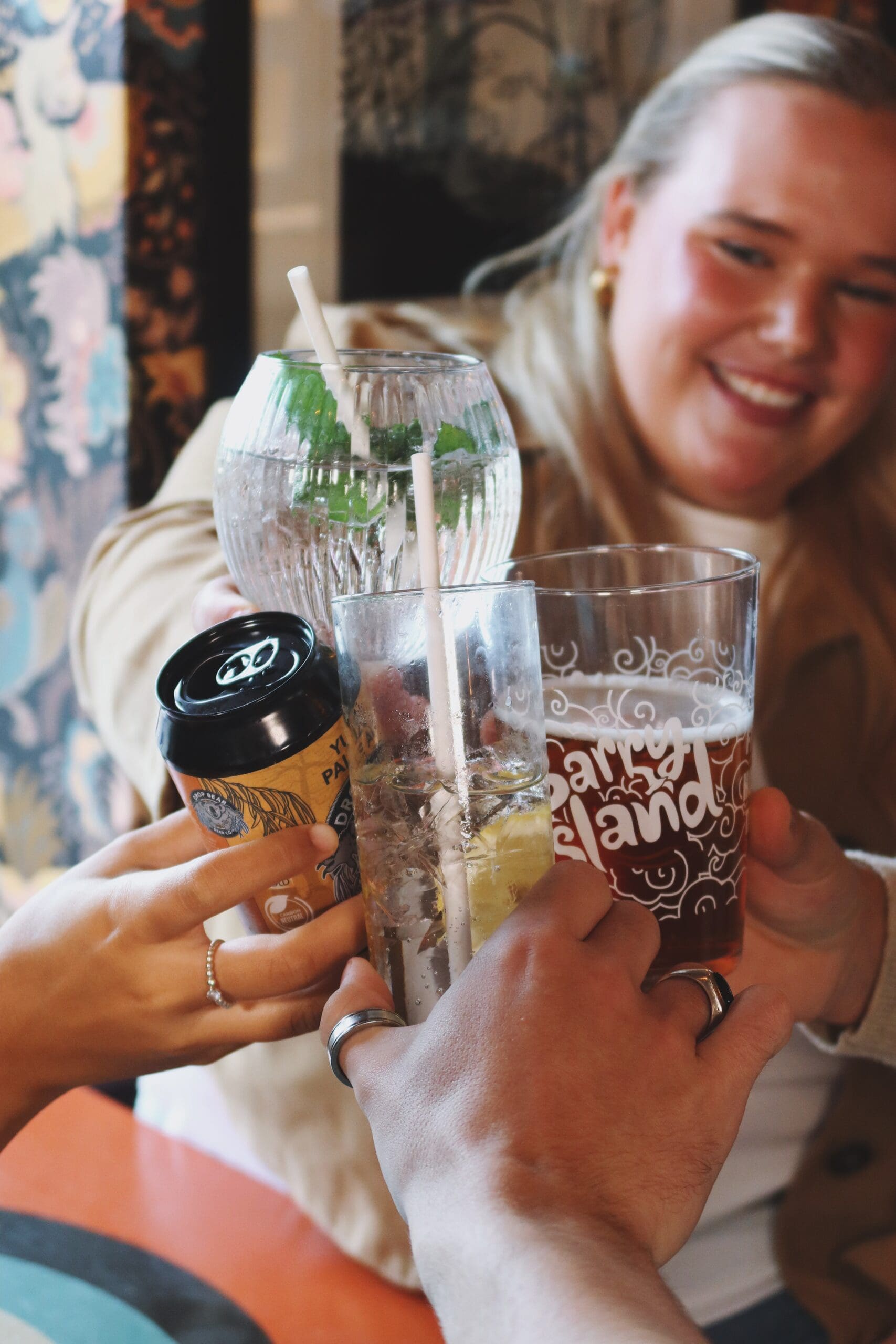 Four people clink various drinks together in a celebratory toast; a smiling person in the background holds a large beer glass while others hold a can, a cocktail, and a glass of water with a straw.