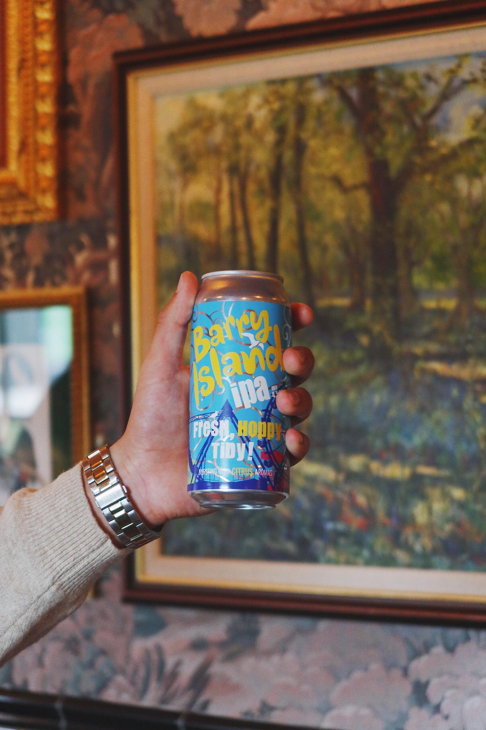 A person wearing a watch holds a can of Barley Island IPA with colorful blue and yellow lettering. A framed painting of a forest hangs on the wall in the background.