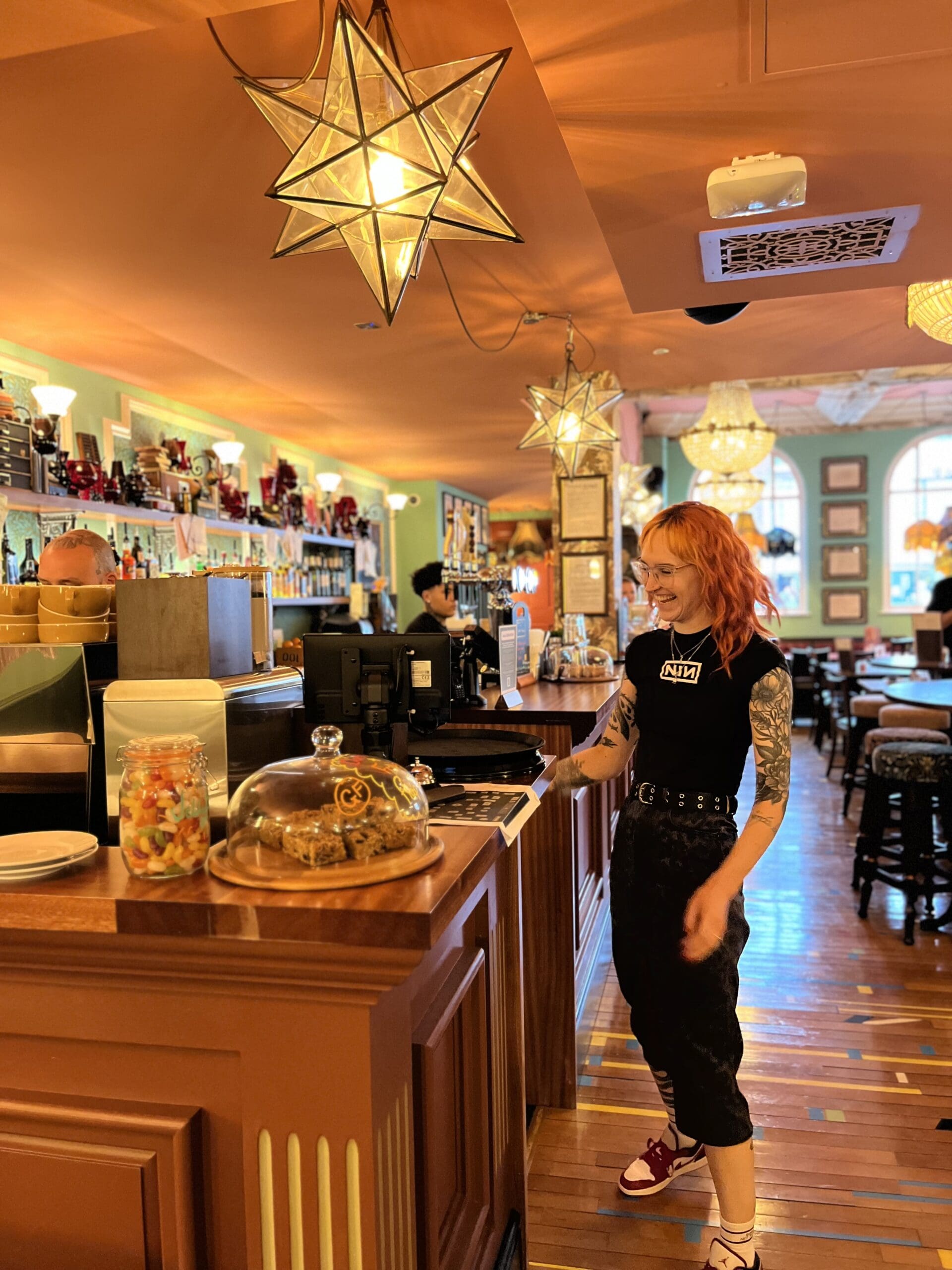A smiling barista with orange hair and tattoos stands behind a wooden counter in a cozy, warmly lit café lounge decorated with star-shaped hanging lamps and shelves filled with jars and bottles.