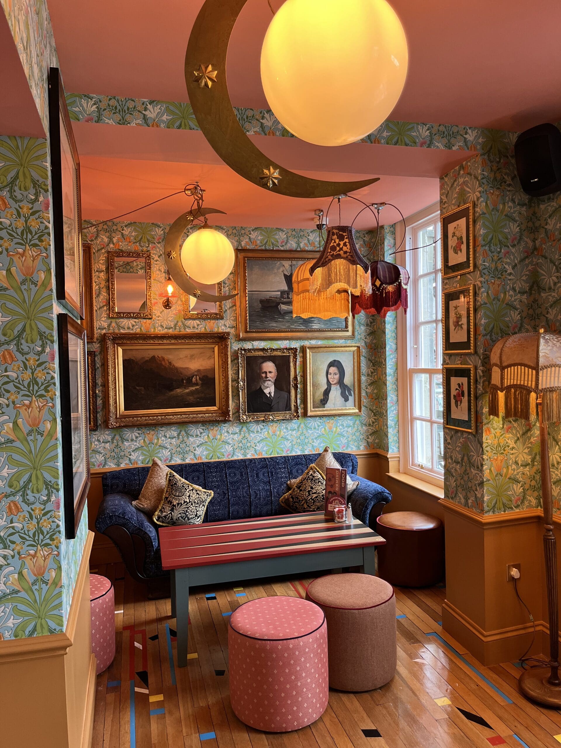 A cozy, colorful lounge with floral wallpaper, framed portraits, patterned cushions, round stools, hanging lamps with tassels, and a crescent moon light fixture creates a whimsical and vintage lounge atmosphere.