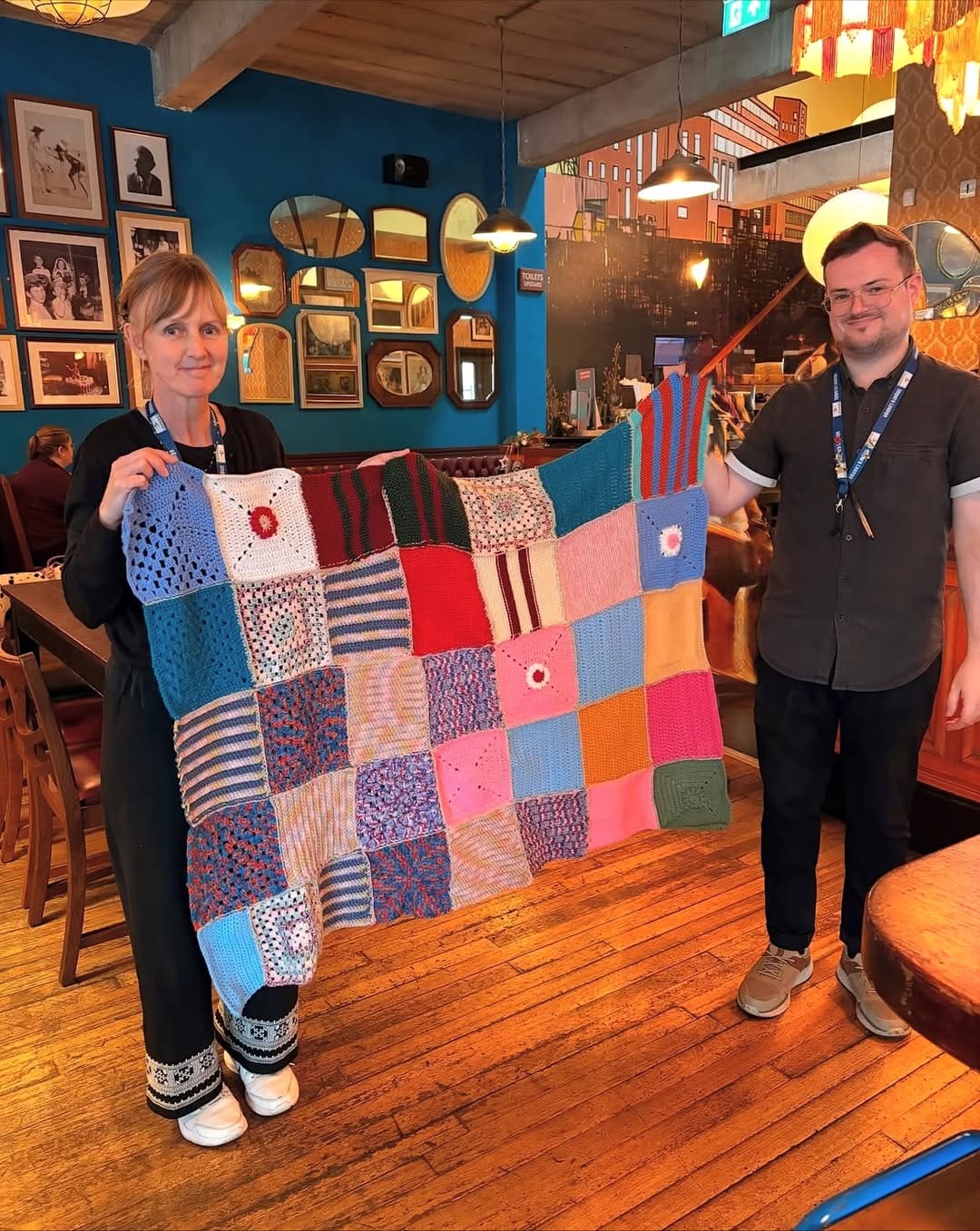 Two people stand in a cozy, colorful café holding a large, patchwork knitted blanket made of various colored and patterned squares. Framed photos and mirrors decorate the walls behind them.