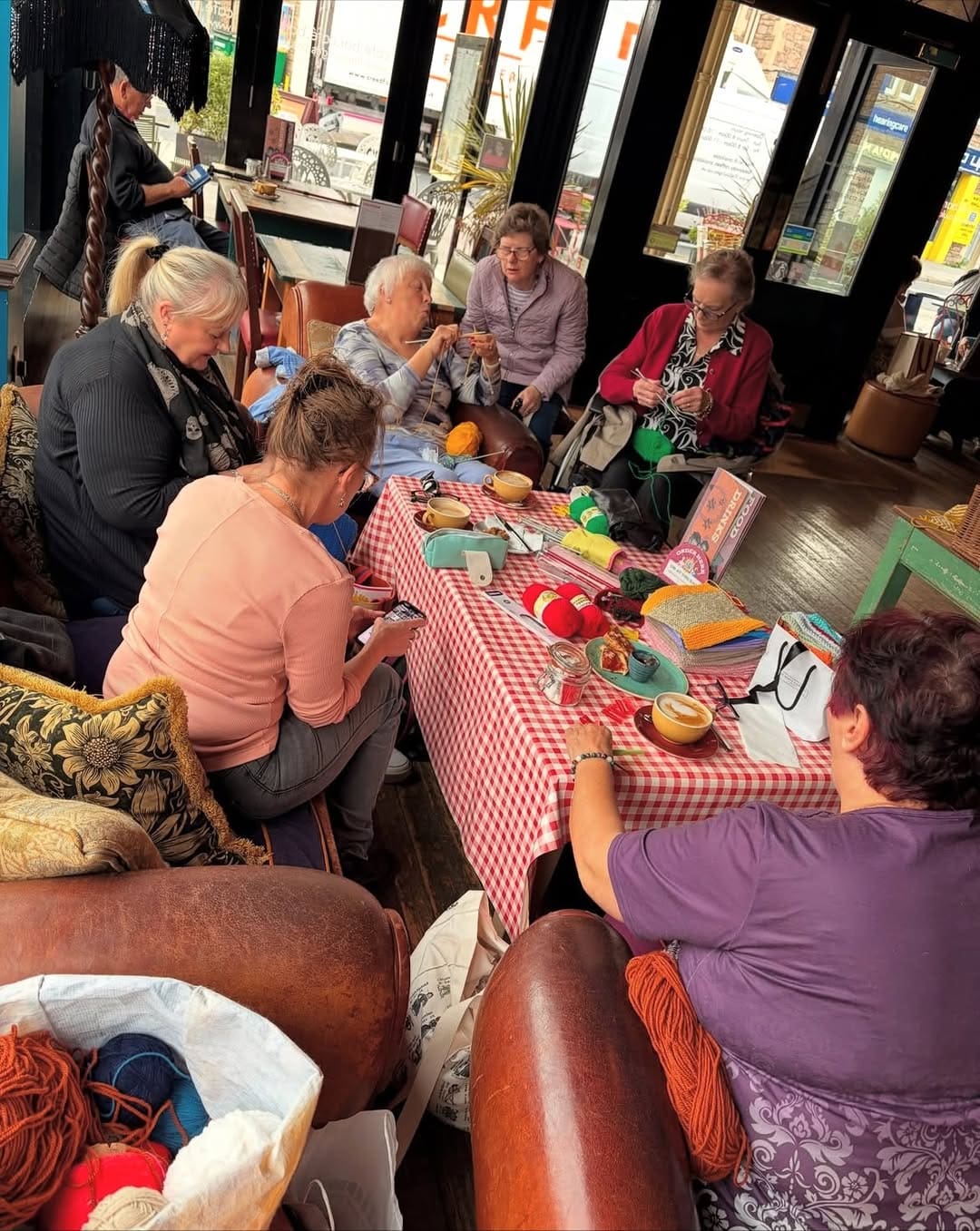 A group of older women sit around a checkered table in a cozy cafe, knitting and chatting. Balls of yarn, knitted items, and supplies are spread out on the table. Large windows let in natural light.