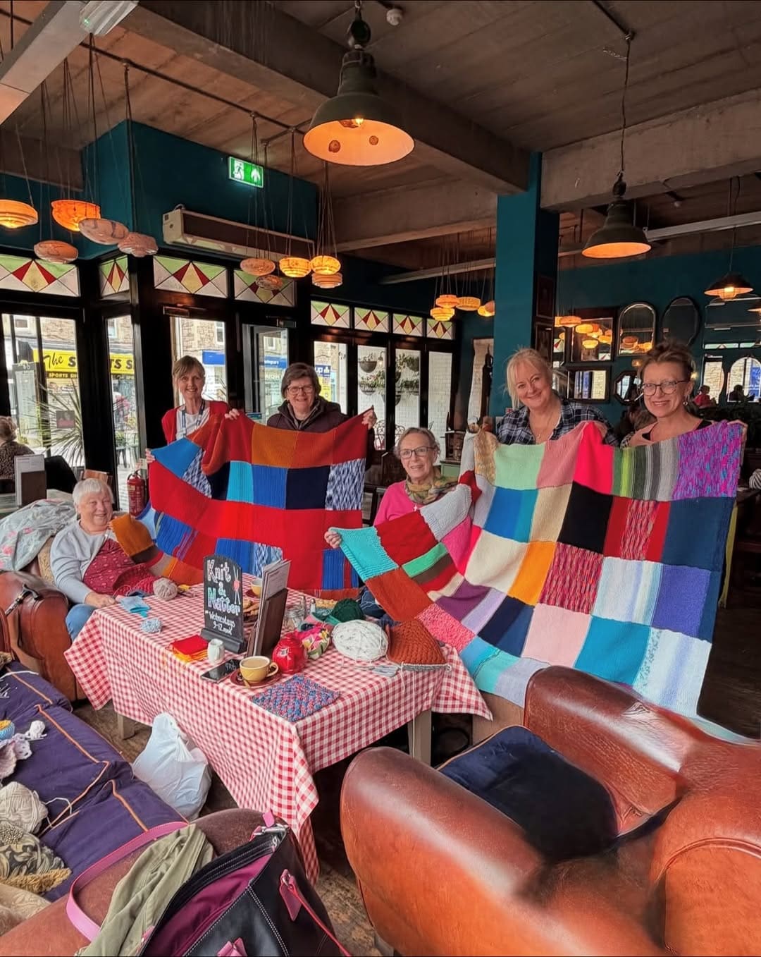 Six women sit and stand around tables in a cozy cafe, smiling and proudly holding up colorful patchwork blankets. Yarn, knitting supplies, and bags are visible on the tables and couches around them. Warm lighting fills the space.